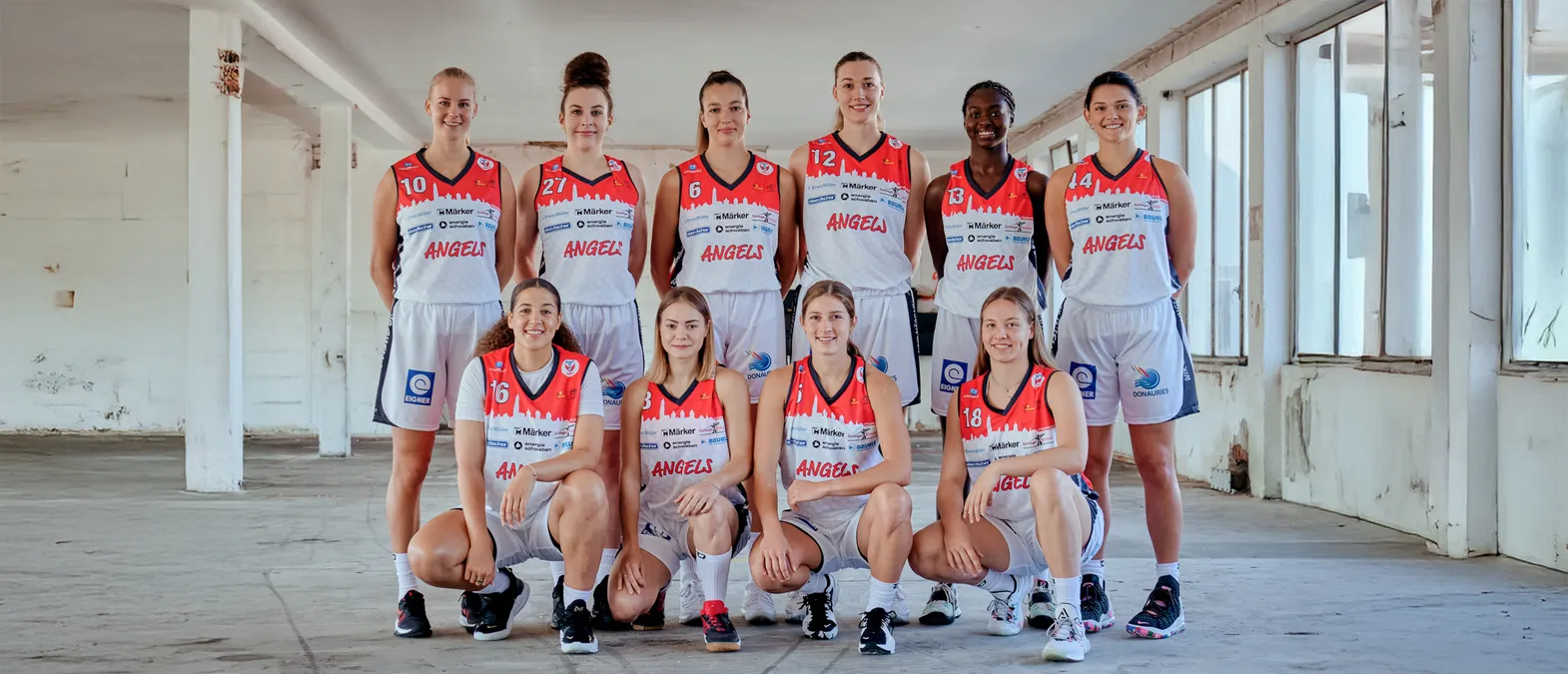 Un groupe de huit femmes portant des maillots de basket-ball orange et blanc avec l'inscription 'ANGELS' posent dans un entrepôt.