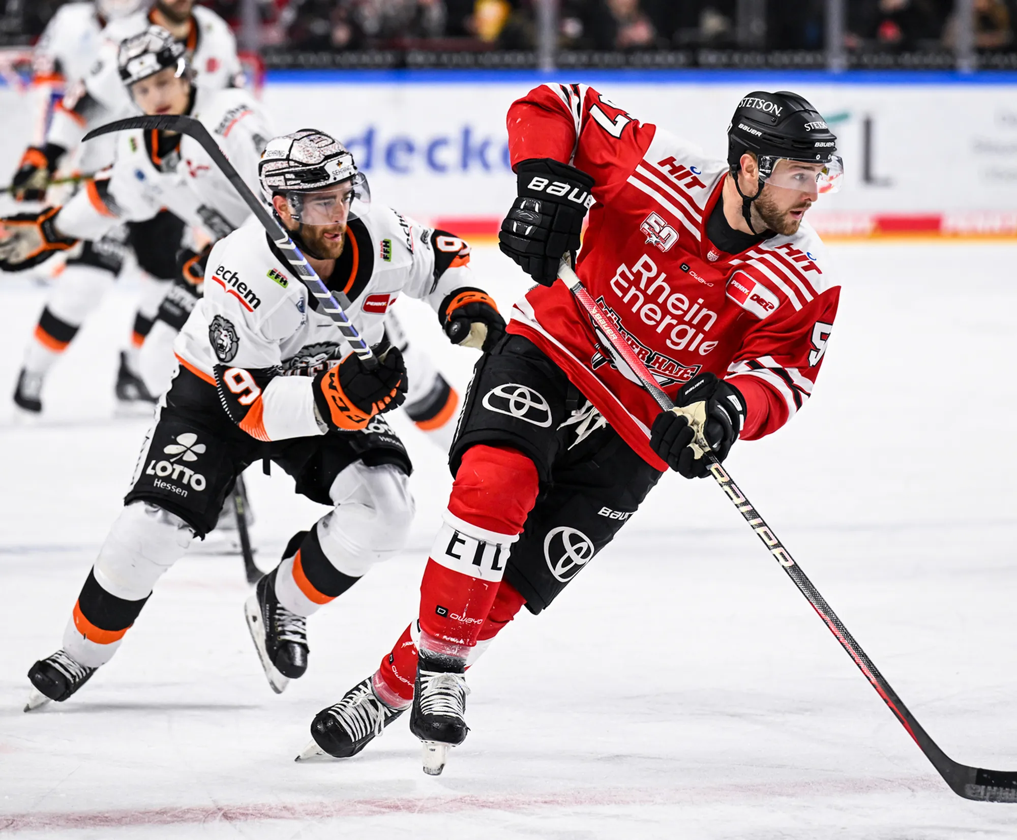 Deux joueurs de hockey sur glace sur la glace, l'un en maillot rouge et l'autre en maillot blanc avec des accents orange.