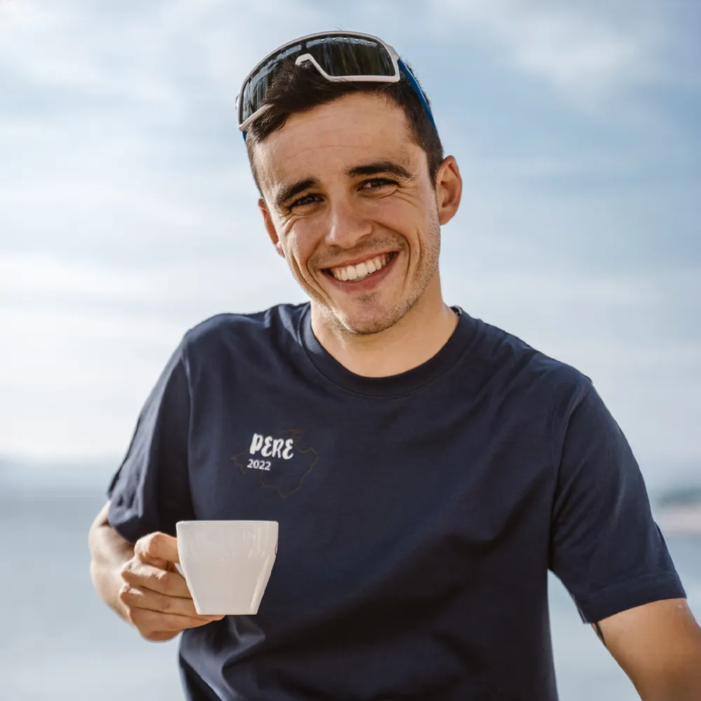 Un homme tient une tasse et porte un t-shirt bleu marine avec l'impression 'PERE 2022', souriant devant un arrière-plan flou.