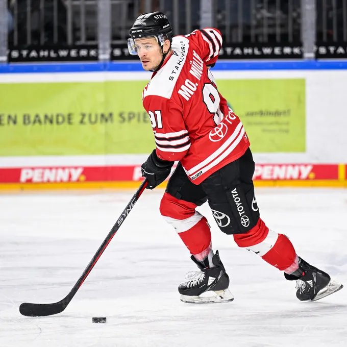 Un joueur de hockey sur glace portant un maillot rouge et noir avec le numéro 91 et le nom Mc. Miller dans le dos, debout sur la glace et contrôlant un palet avec sa crosse.