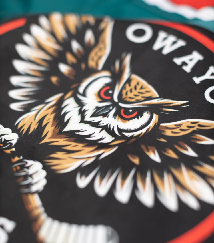 Un gros plan d'un maillot de sport noir avec un logo d'hibou stylisé tenant une crosse de hockey, et le texte 'owayo' en lettres blanches.
