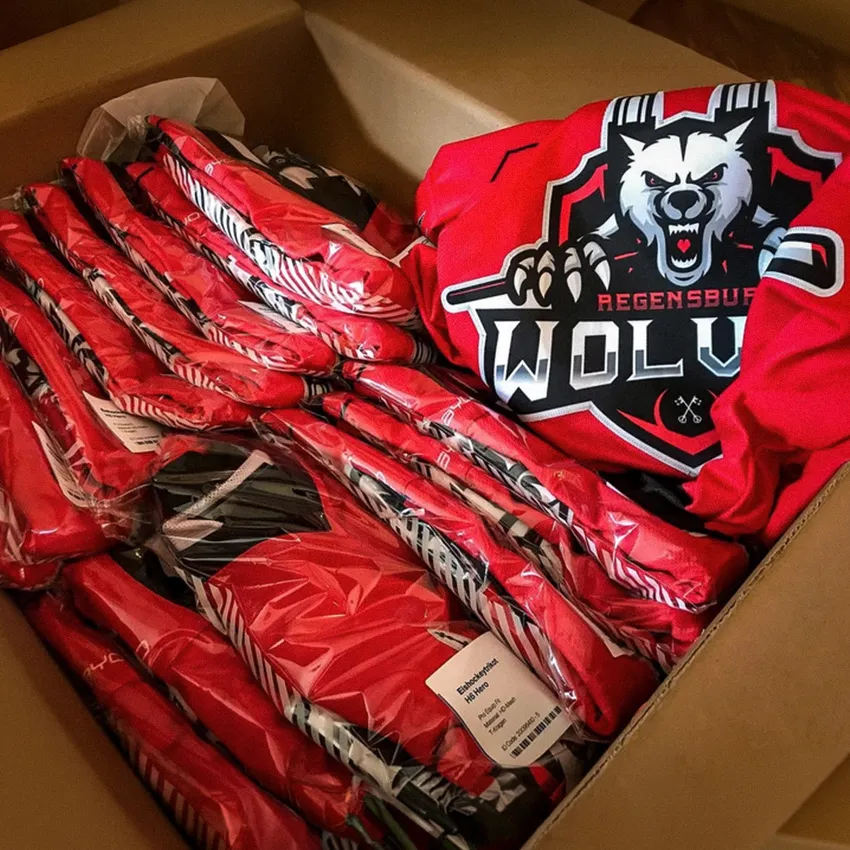 Une boîte remplie de plusieurs maillots de sport rouges emballés sous plastique, avec un maillot visible arborant un logo de loup et le texte 'Regensburg WOLV'.