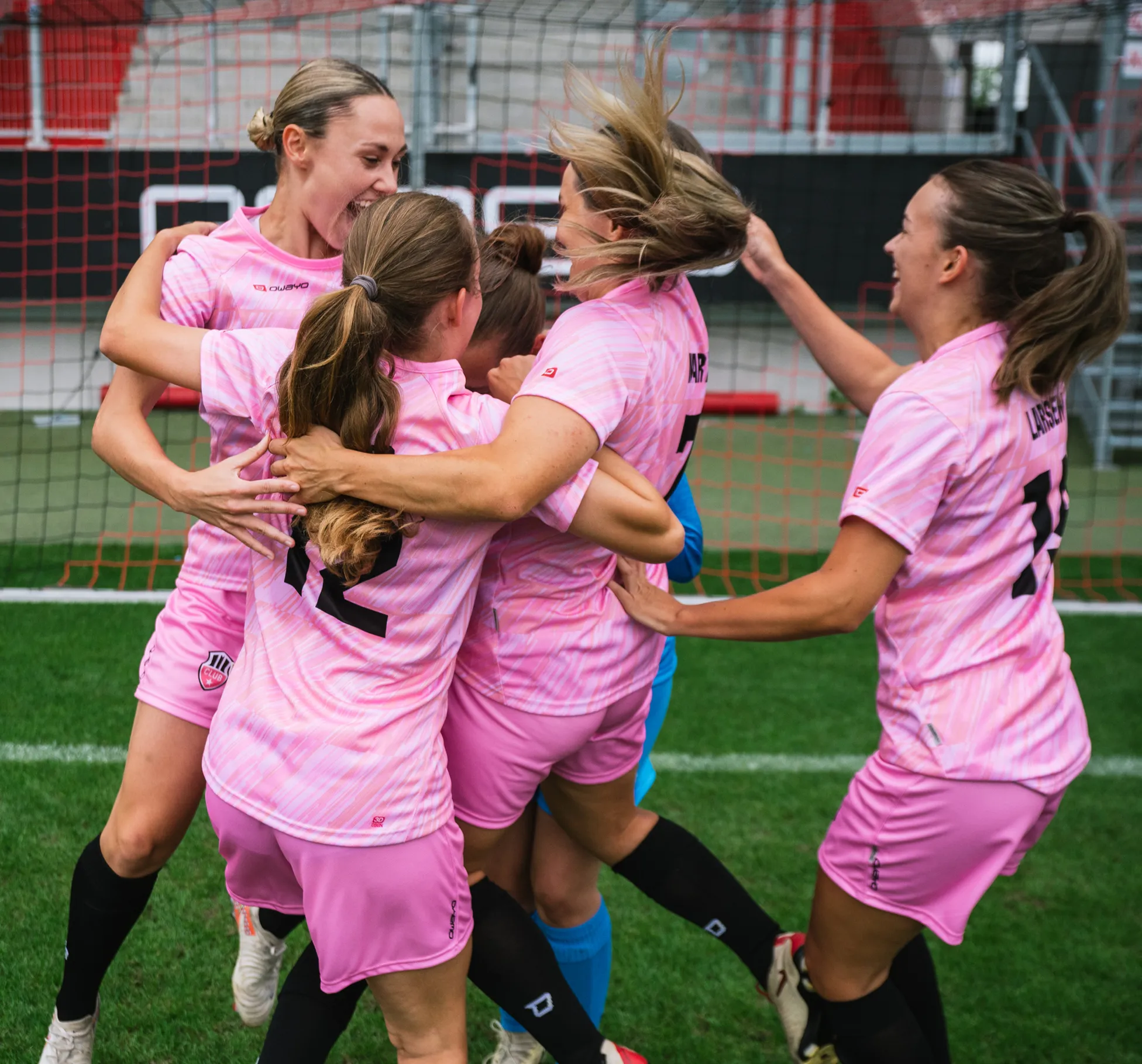 Cinq joueuses en maillots roses célèbrent joyeusement ensemble sur le terrain de football.