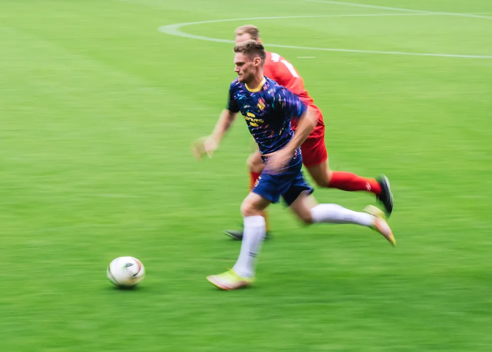 Un joueur de football en maillot bleu avec des motifs colorés court rapidement sur le terrain vert tandis qu'un autre joueur le suit de près.