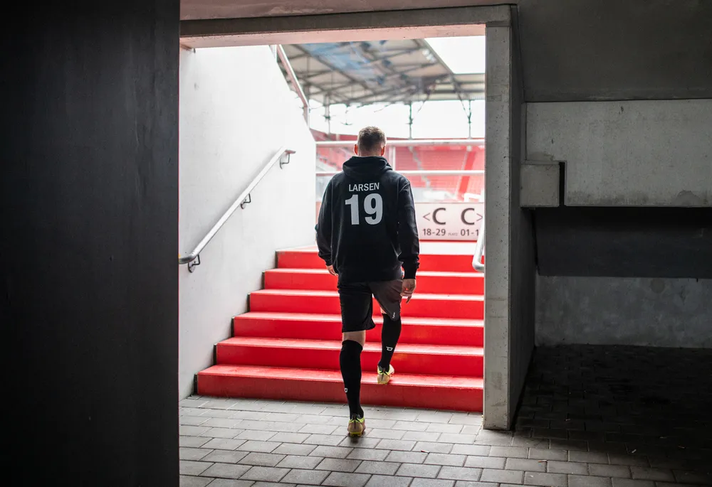 Un joueur de football portant un sweat à capuche noir avec le numéro 19 et un short monte les marches rouges d'un stade.