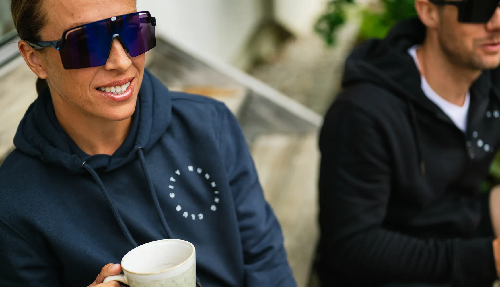 Une personne en sweat bleu tient une tasse et porte de grandes lunettes de soleil, tandis qu'une autre personne en arrière-plan porte également un sweat.