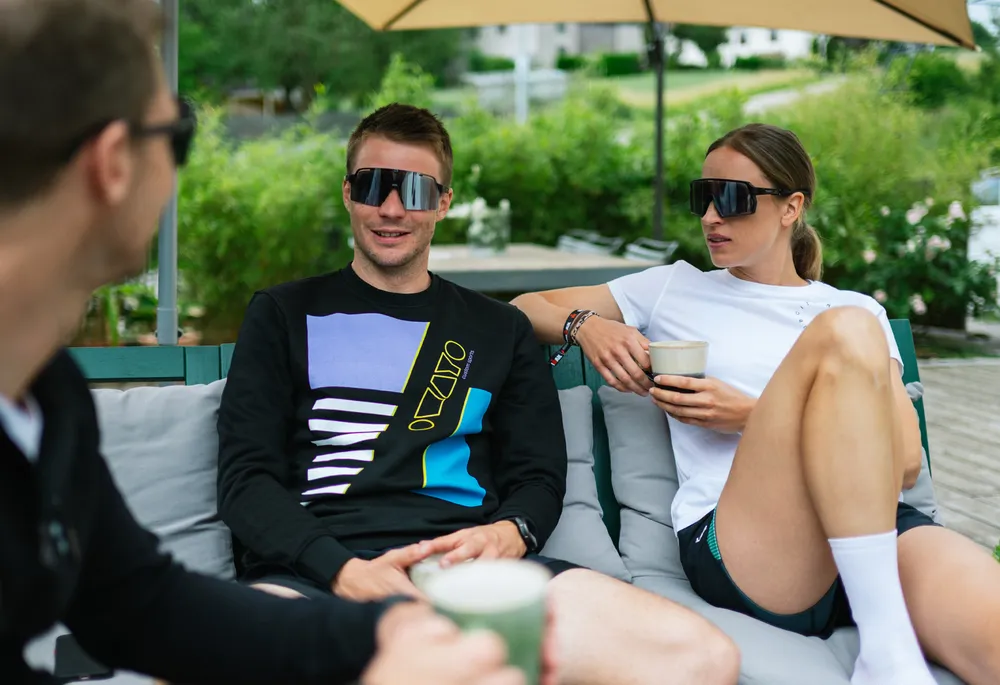 Trois personnes sur un canapé, deux portant des vêtements de sport décontractés et des lunettes de soleil, tandis qu'elles se détendent et discutent en tenant des boissons.