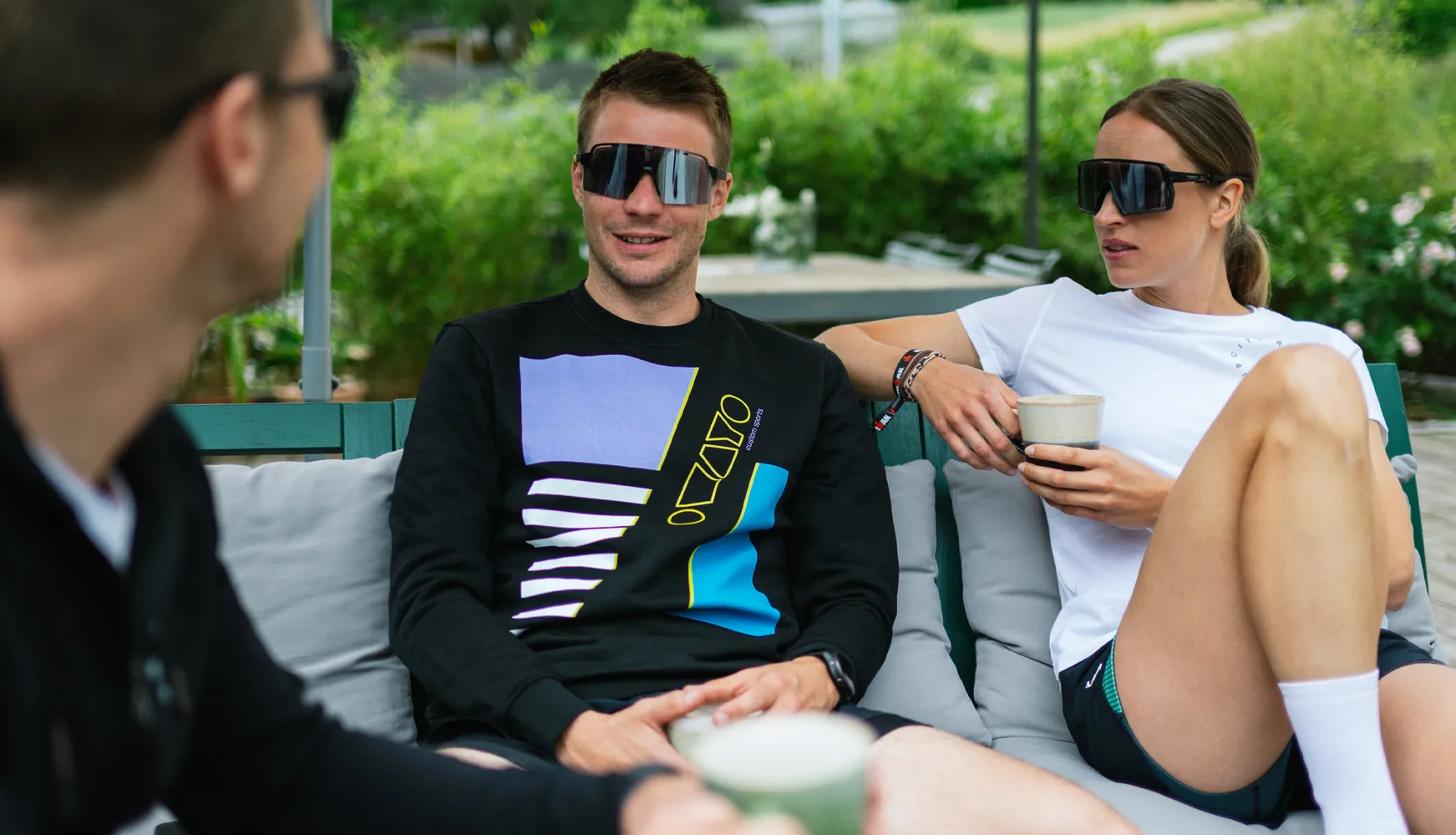 Trois personnes sont détendues sur un canapé, l'une portant un sweat noir avec un design coloré et l'autre un t-shirt blanc, toutes deux tenant des boissons.