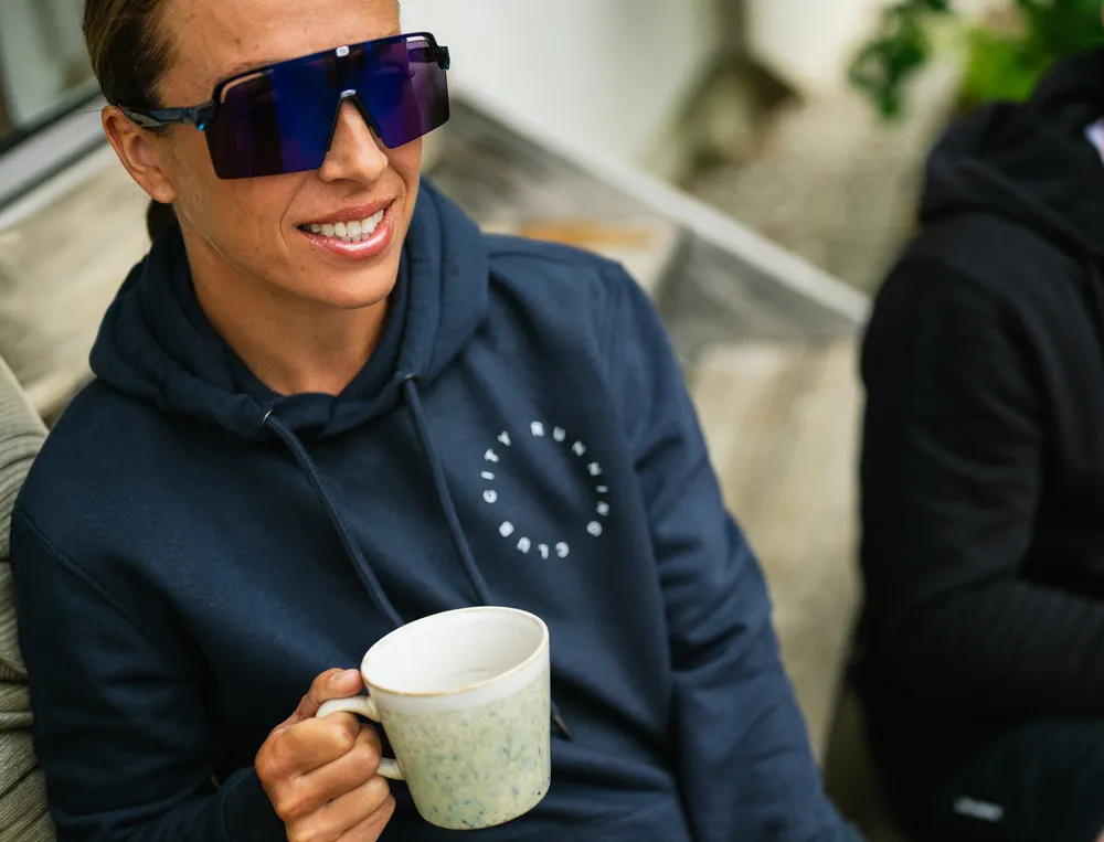 Une personne portant un hoodie bleu marine avec un logo tient une tasse de café et porte de grandes lunettes de soleil.