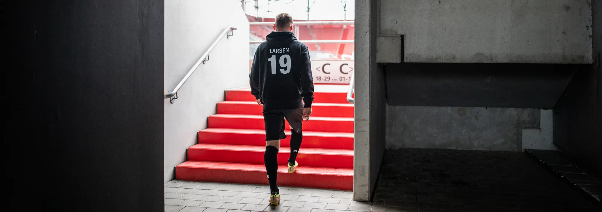 Un joueur en sweat à capuche noir et en short monte des escaliers rouges sortant d'un stade.