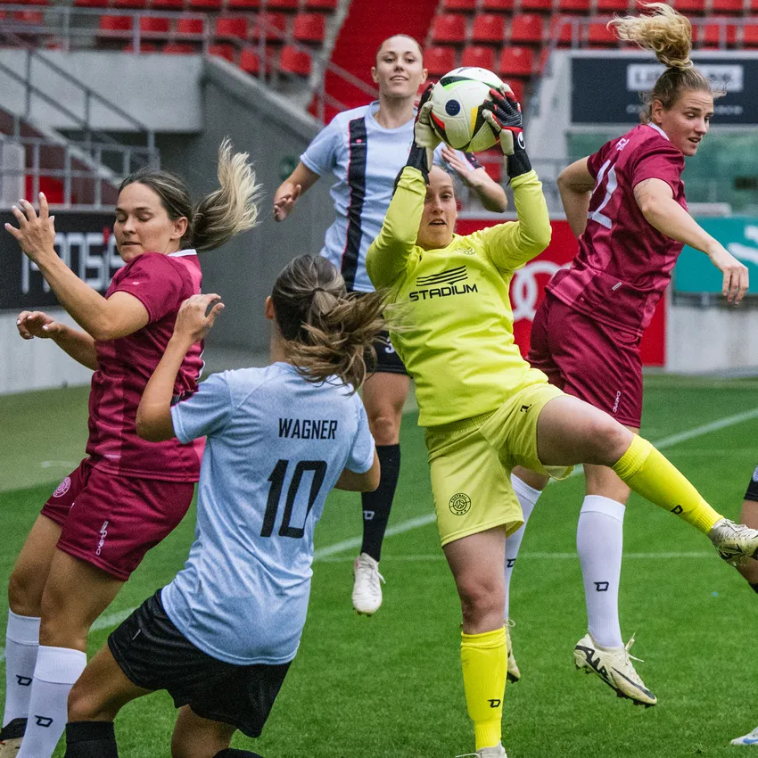 Une gardienne de but en maillot jaune attrape le ballon pendant que des joueuses en maillots variés sont actives sur le terrain de football.