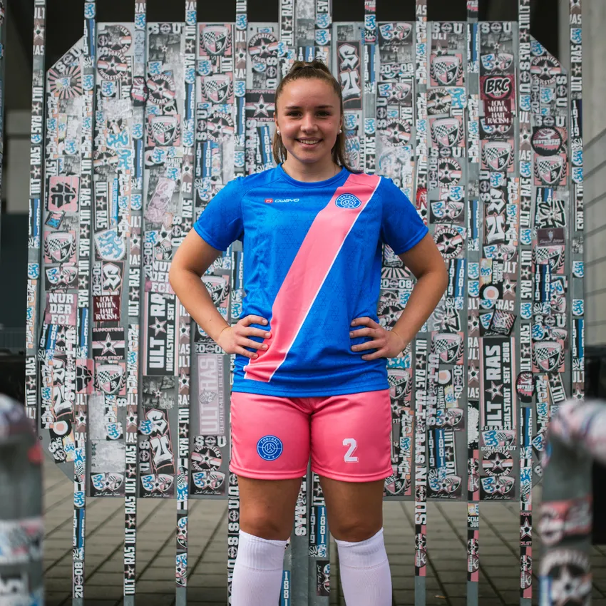 Une fille sourit devant un mur recouvert d'autocollants, portant un maillot de football bleu avec une rayure rose et un short rose.