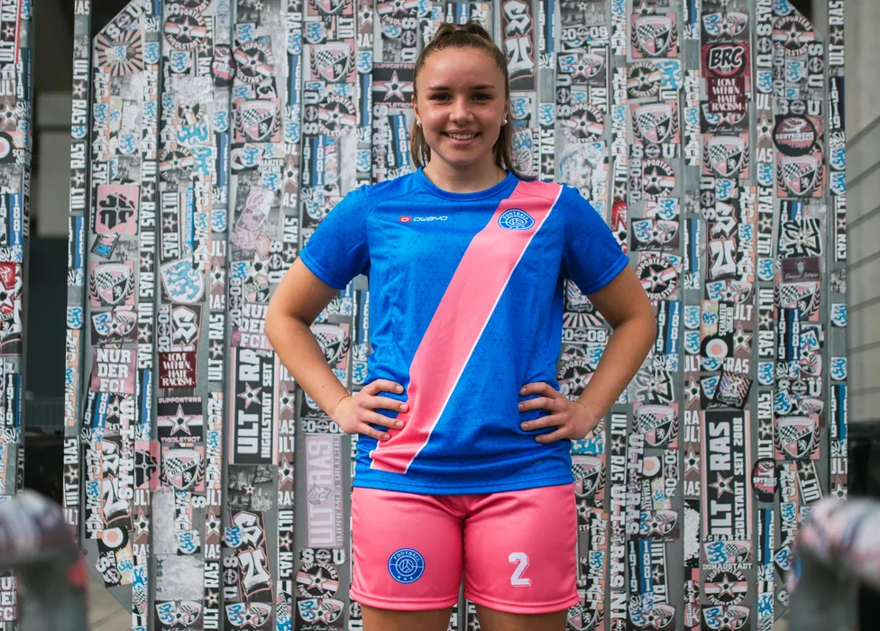Une jeune athlète pose dans un maillot de football bleu avec une bande rose et un short rose devant un mur coloré couvert d'autocollants.