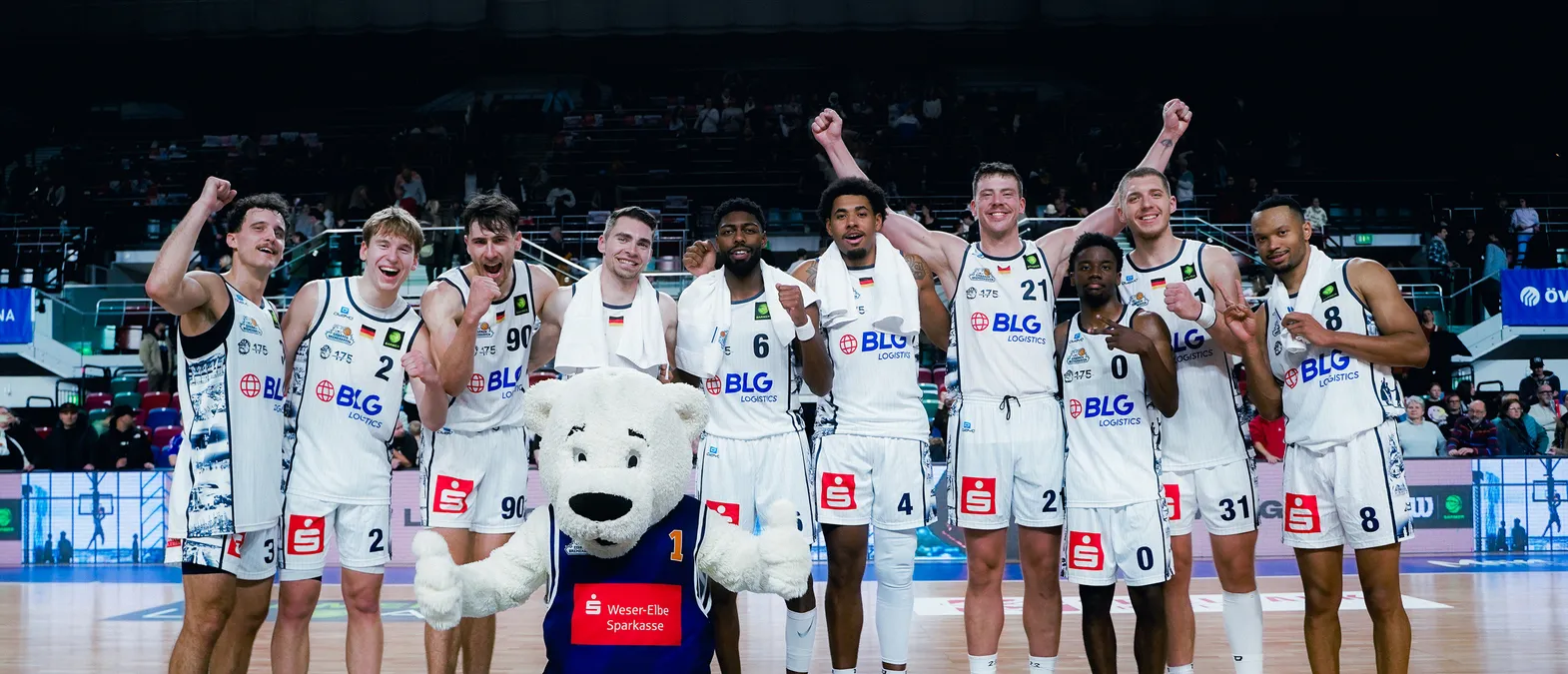 Des joueurs de basket-ball en maillots blancs avec le logo de BLG Logistics célèbrent sur le terrain, entourés d'un public, avec un mascotte ours polaire au premier plan.