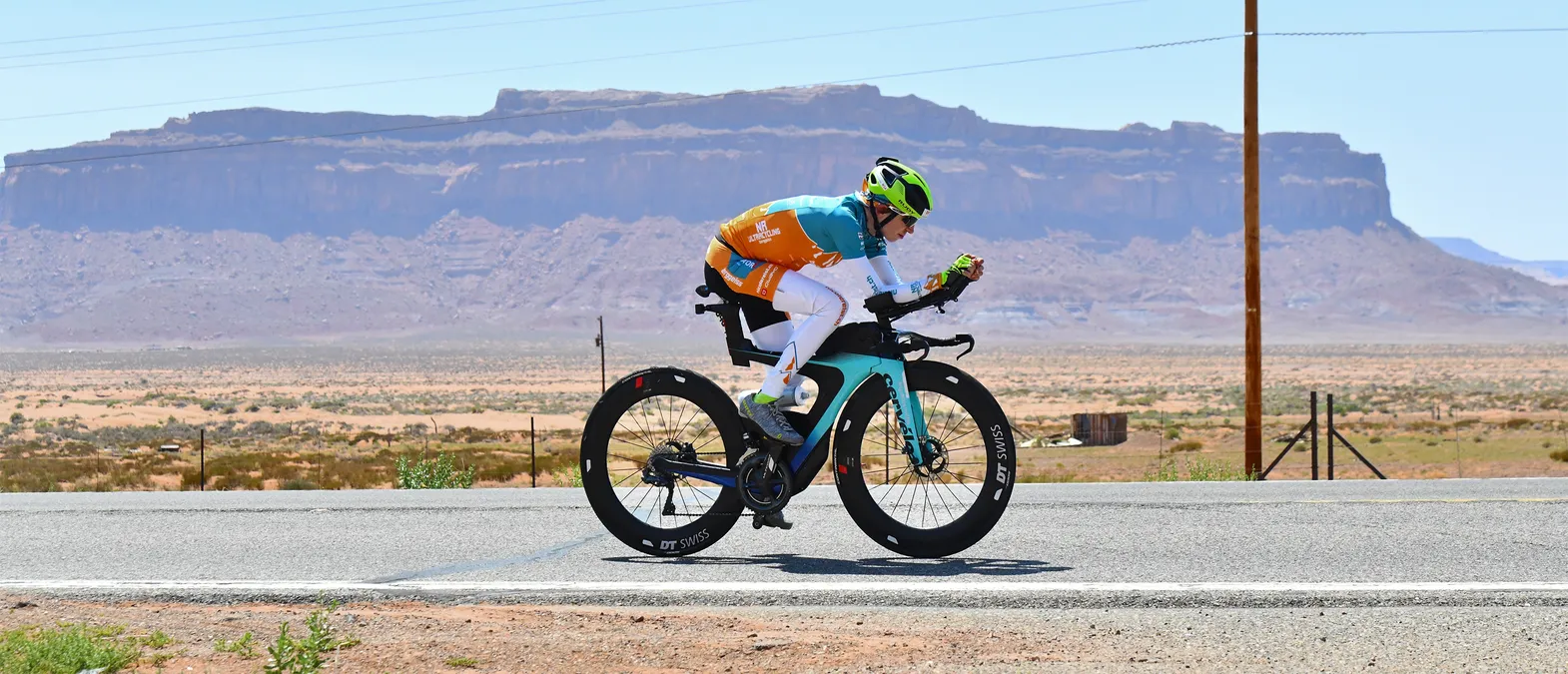 Une cycliste en maillot orange et turquoise pédalant sur un vélo de contre-la-montre sur une route dans un paysage désertique avec des formations rocheuses en arrière-plan.