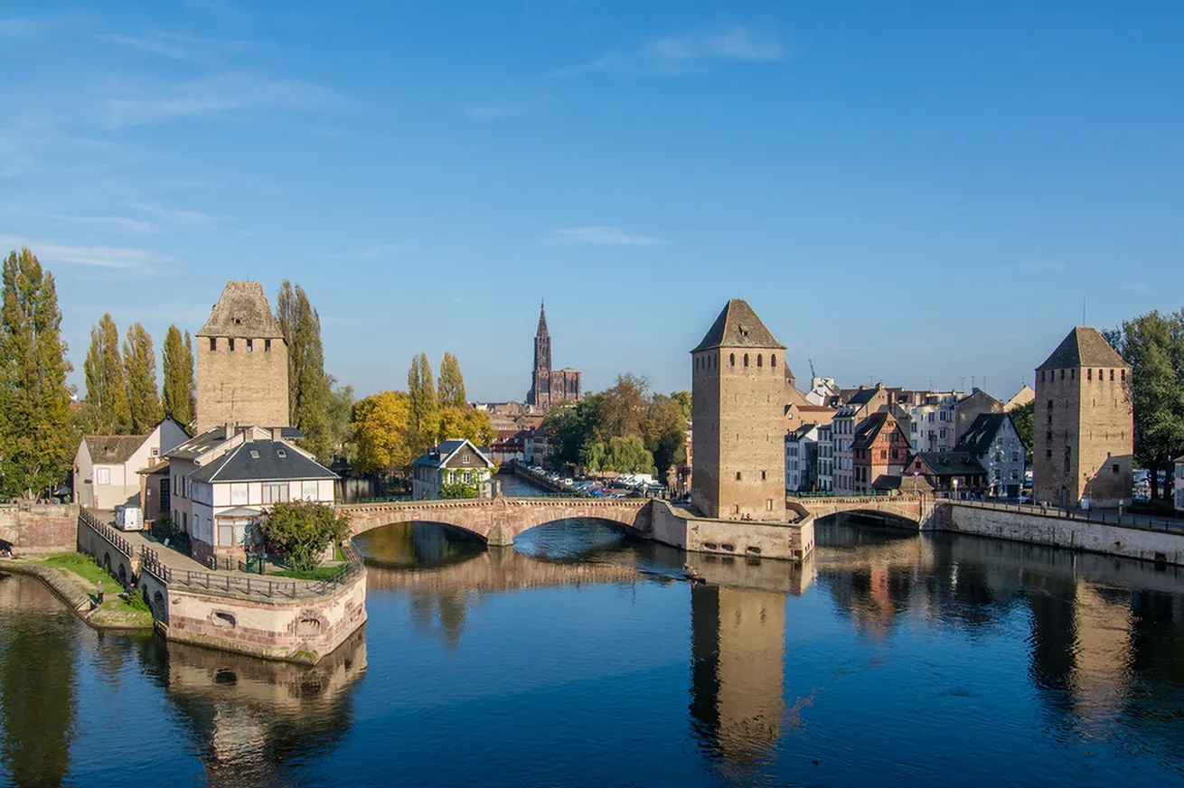 Une vue de la vieille ville de Strasbourg avec les tours des Ponts Couverts au-dessus de la rivière, sous un ciel bleu.