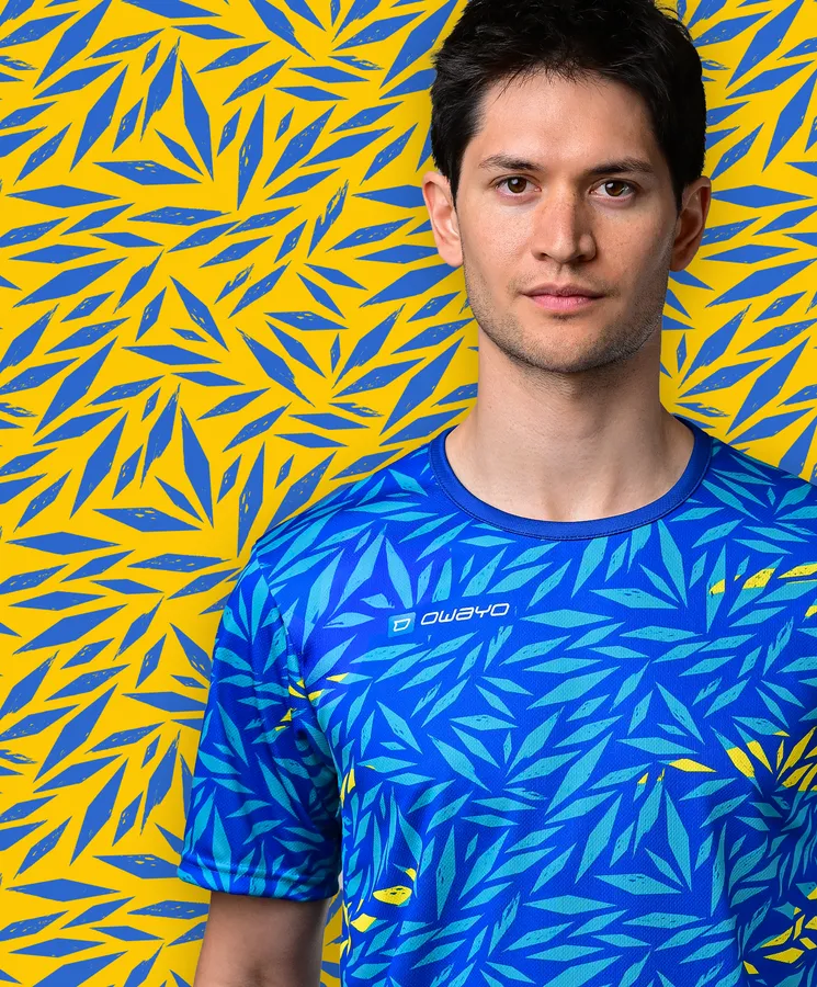 Un jeune homme porte un T-shirt de sport bleu avec un motif de feuilles colorées sur un fond jaune.