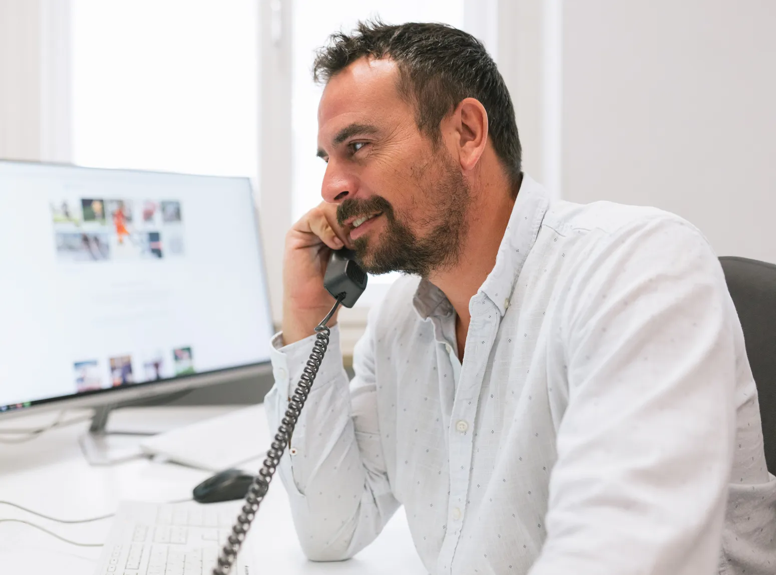 Un homme barbu est assis à un bureau devant un ordinateur, souriant tout en parlant sur un combiné téléphonique à l'ancienne.