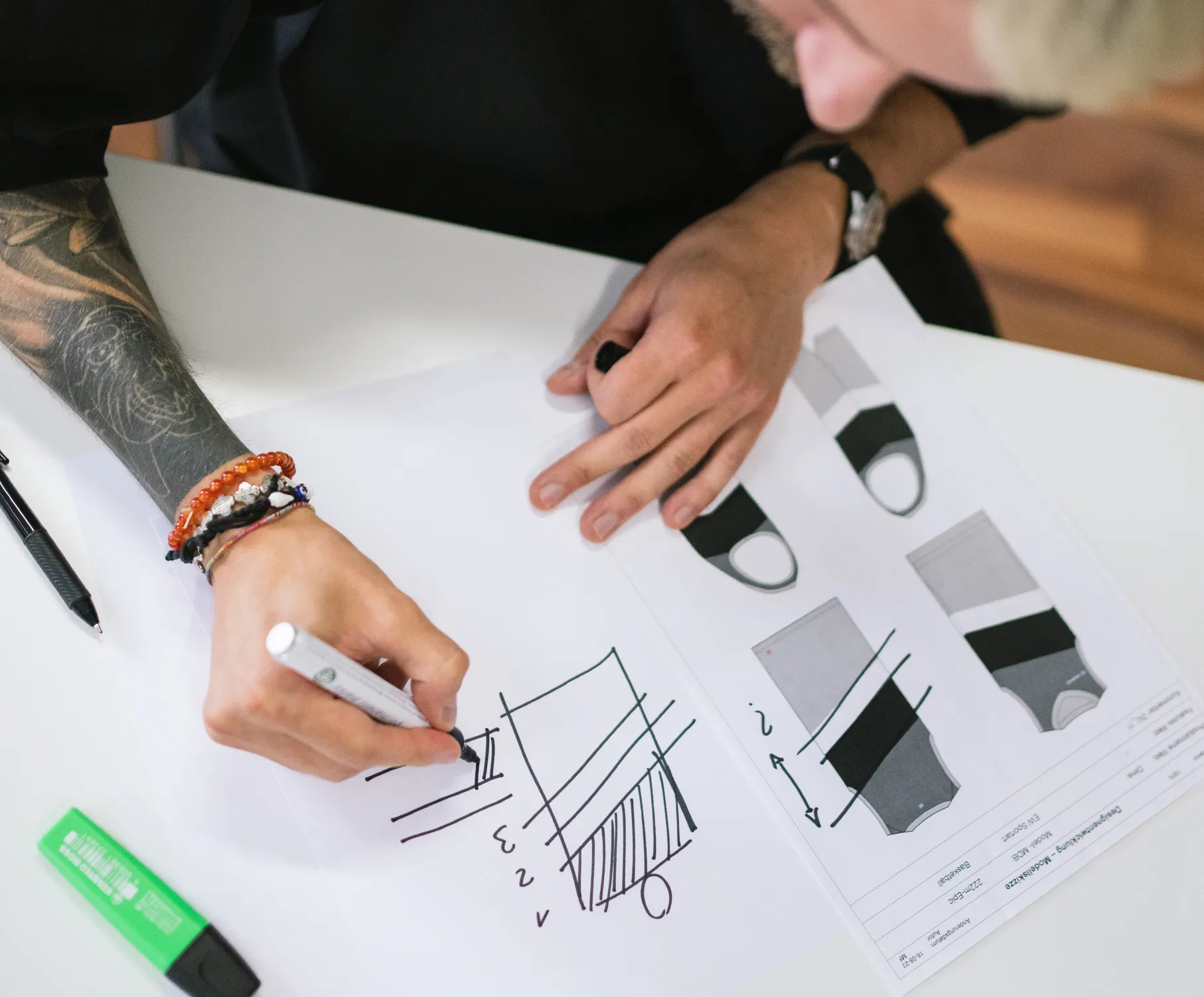 Une personne avec un tatouage au bras dessine un design sur du papier avec un marqueur, tandis que des dessins techniques de vêtements de sport sont posés sur la table.
