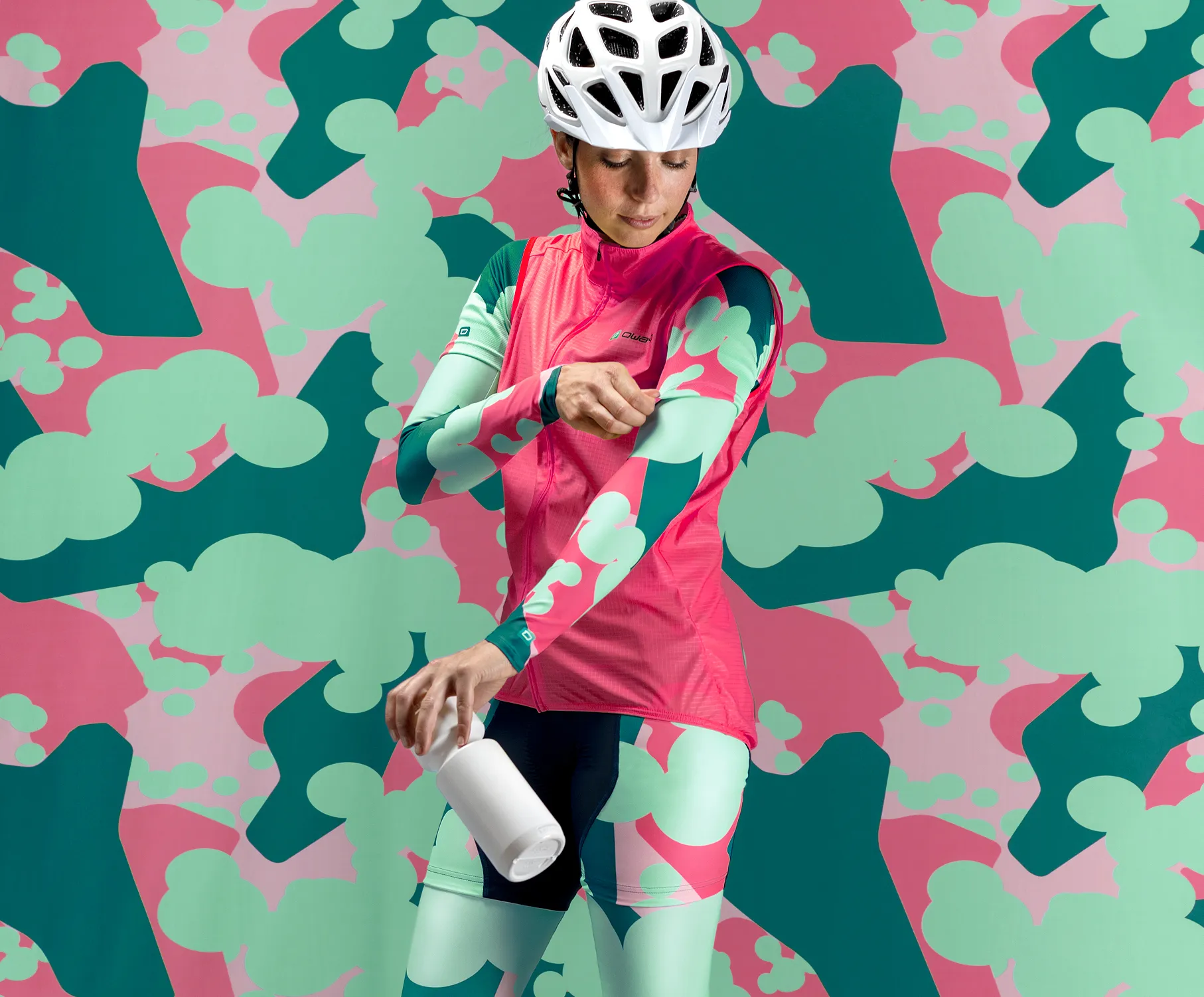 Une cycliste portant une tenue de sport personnalisée rose et verte avec un motif abstrait, ajustant ses manches tout en tenant une bouteille blanche, devant un fond coloré avec le même motif.