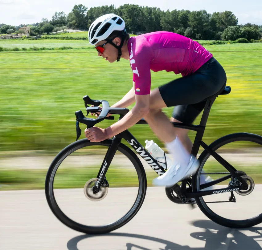 Un cycliste en maillot rose et short noir roule sur une route à travers un paysage verdoyant, la floue de mouvement soulignant la vitesse.