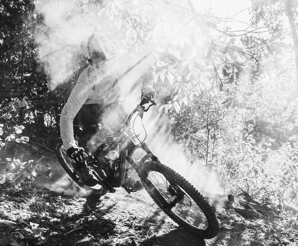 Un cycliste de montagne en mouvement sur un sentier forestier, entouré de poussière et de rayons de lumière, portant un haut à manches longues d'owayo.