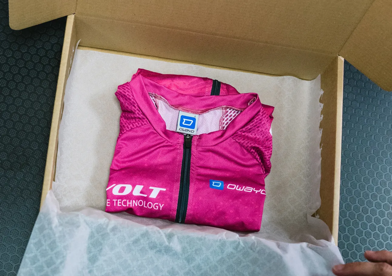 Un maillot de sport sans manches rose plié avec le logo owayo est placé dans une boîte en carton sur du papier blanc.