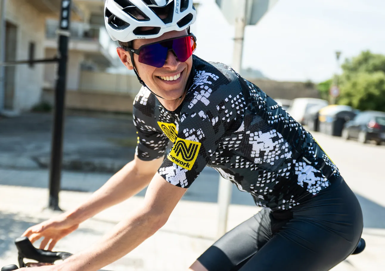 Un cycliste portant un maillot de cyclisme noir et blanc à motif avec un logo sponsor jaune, roulant dans la rue.