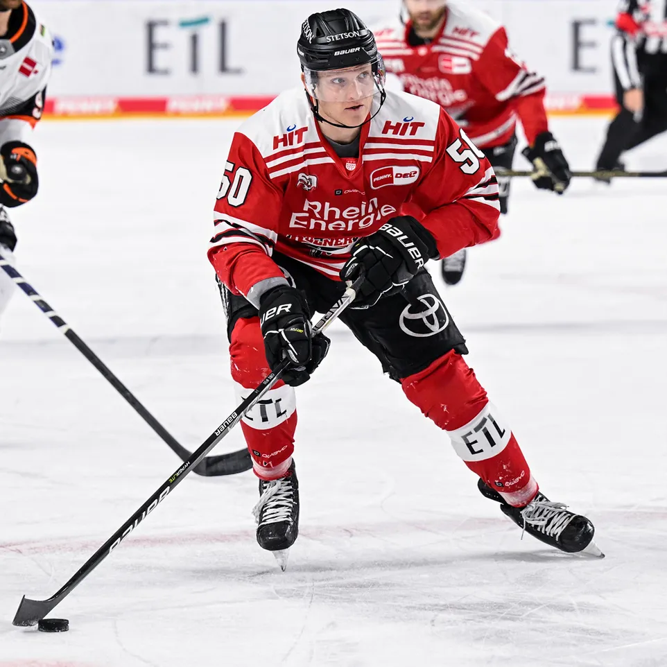 Un joueur de hockey sur glace portant un maillot rouge avec le numéro 50 et l'inscription 'Rhein Energie', sur la glace, contrôlant un palet avec sa crosse.