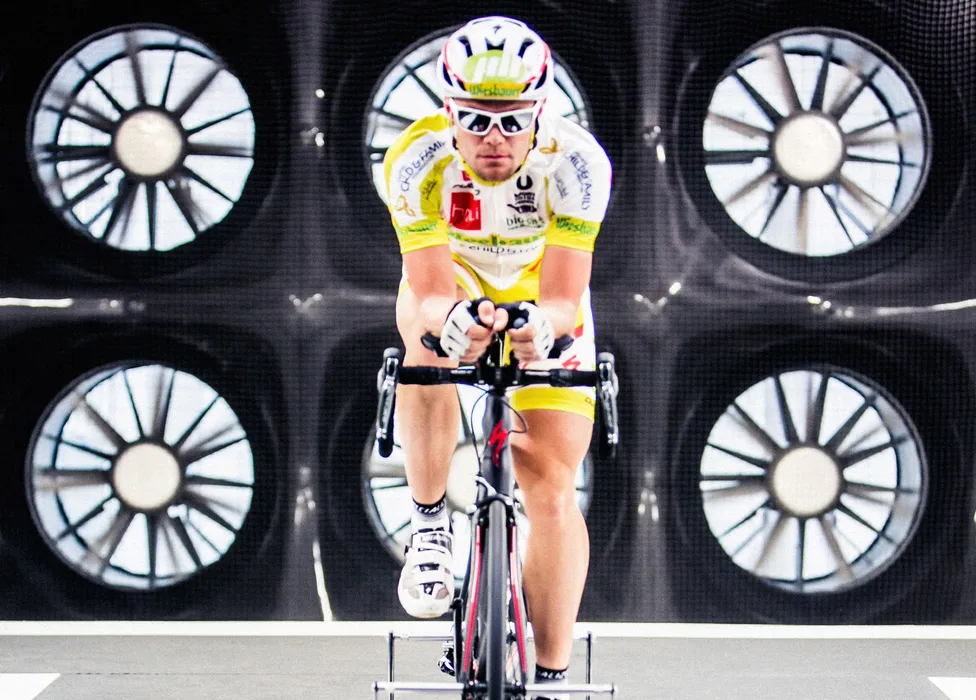 Un cycliste en maillot jaune et blanc et casque pédale sur un vélo d'entraînement devant un fond avec de grands ventilateurs.