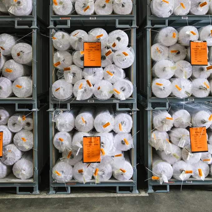 Un entrepôt avec des étagères remplies de grandes rouleaux de tissu blanc emballés dans du plastique et portant des étiquettes orange.
