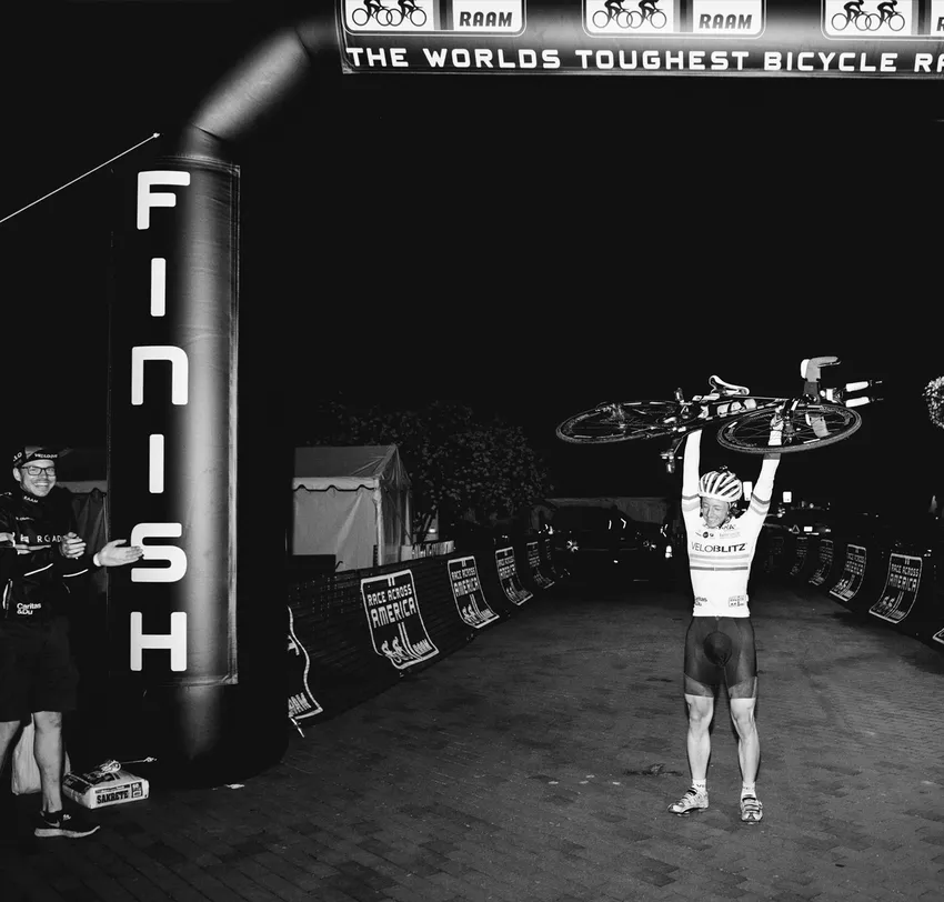 Un cycliste célèbre sa victoire en franchissant la ligne d'arrivée de la 'Race Across America' en soulevant son vélo au-dessus de sa tête, tandis qu'un homme l'applaudit à côté.