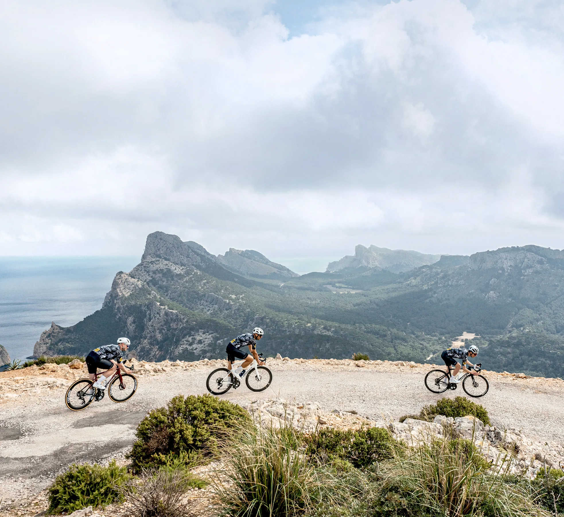 Trois cyclistes en vêtements de sport roulent sur une route le long d'un paysage côtier avec des montagnes et la mer en arrière-plan.
